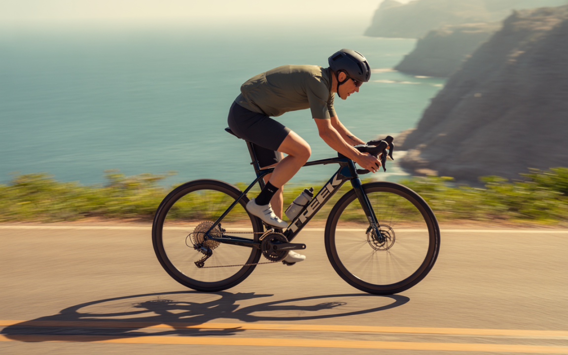 Un cycliste en tenue de sport roule rapidement sur une route côtière, avec l'océan et des collines en arrière-plan.