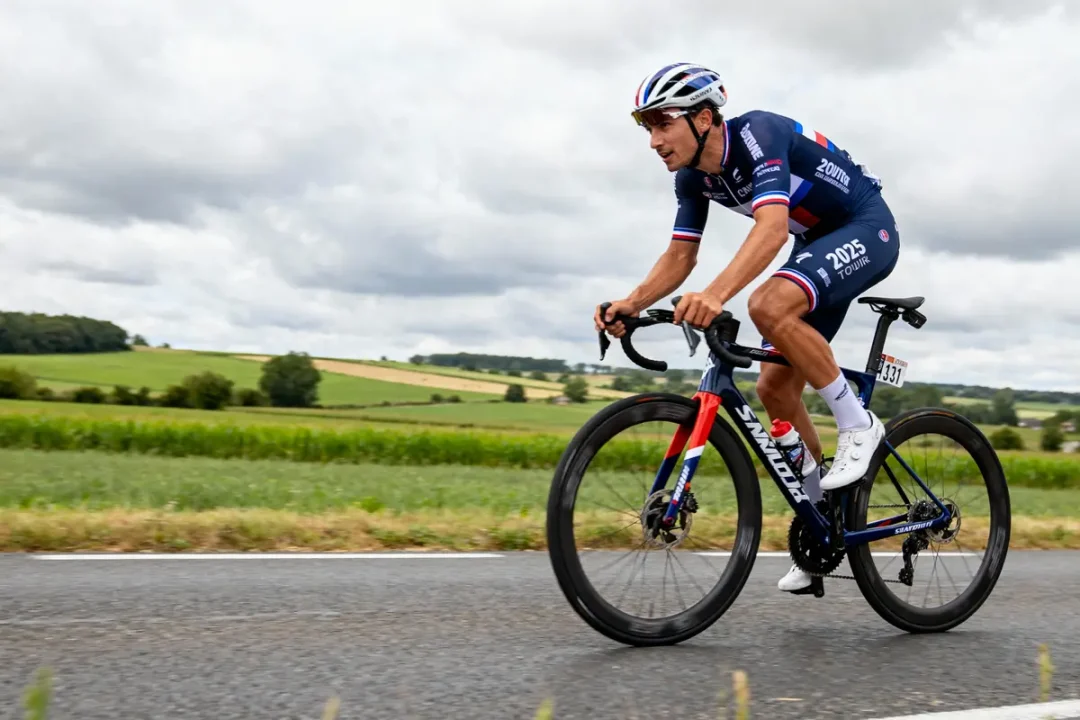 Cycliste en compétition sur route, portant un maillot bleu, paysage de campagne en arrière-plan, ciel nuageux.