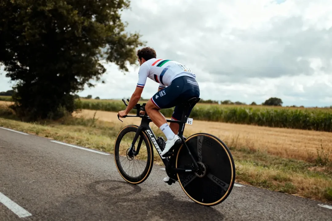Cycliste sur route de campagne, vêtu d'un maillot blanc, chevauchant un vélo noir avec roues aérodynamiques.