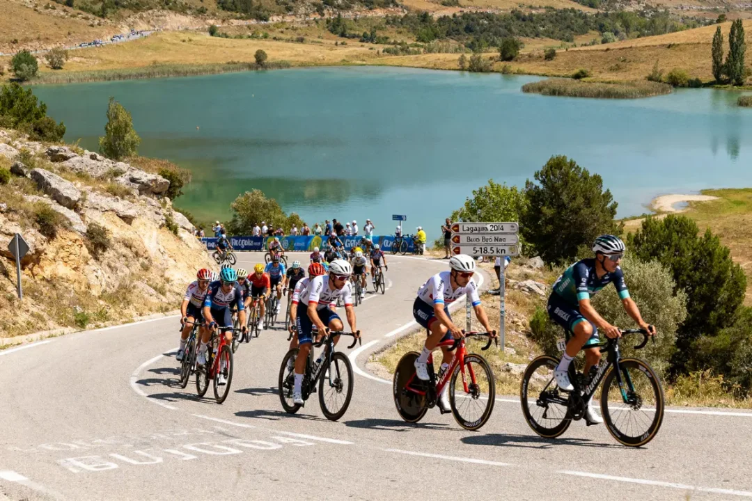 Cyclistes en course près d'un lac, paysage montagneux en arrière-plan, route sinueuse, signalisation routière.