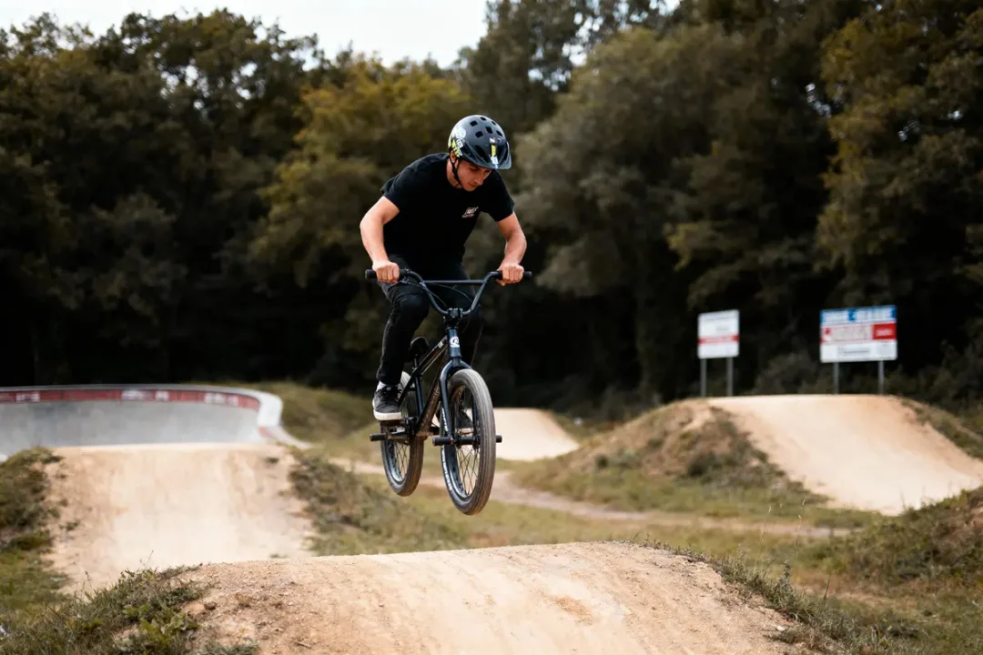 Jeune homme en BMX effectuant un saut sur une piste de terre, entourée de forêt.