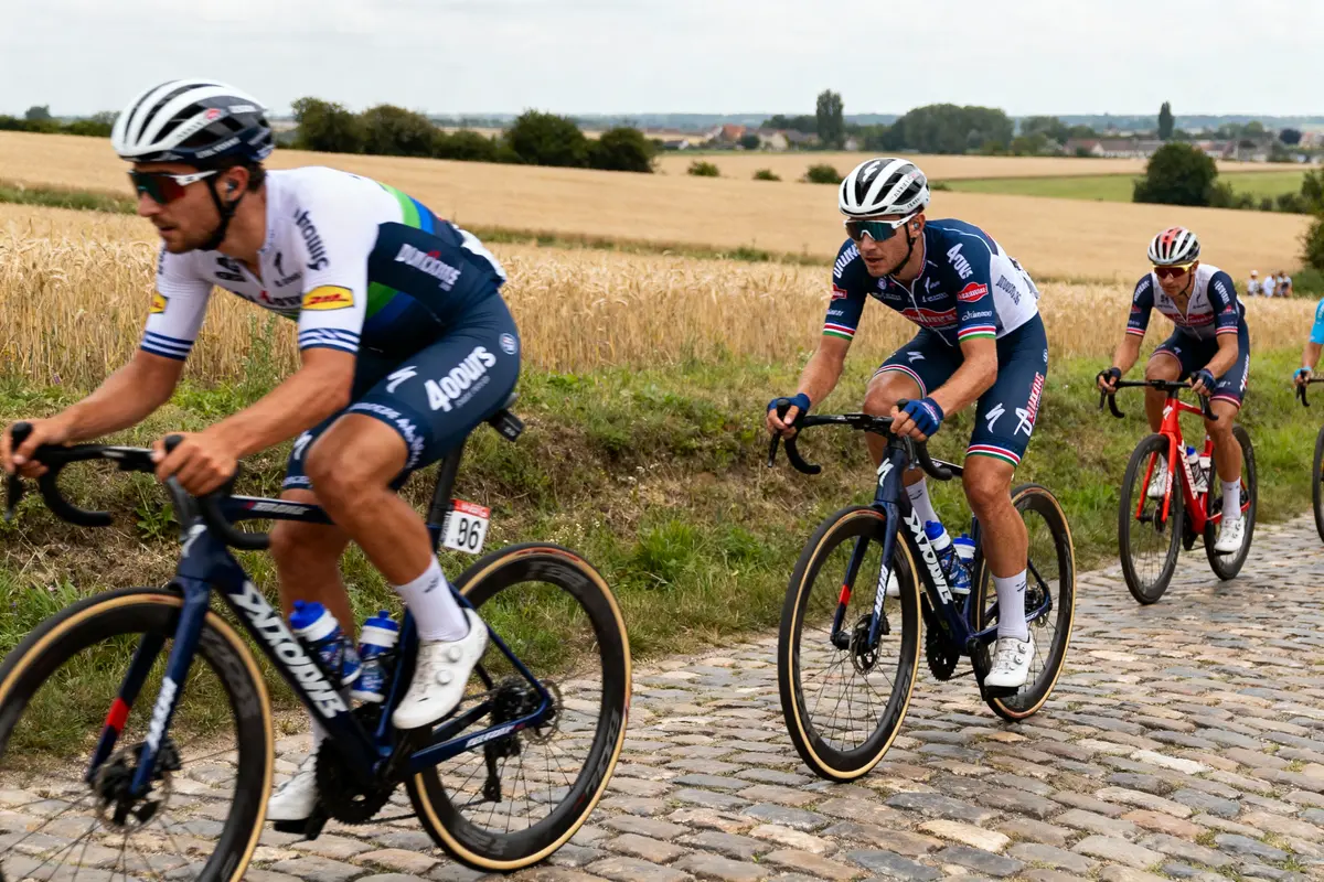 Cyclistes sur route pavée, paysage rural, ciel légèrement nuageux, cyclisme de course, trois cyclistes en tête.