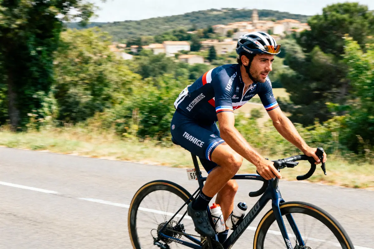 Cycliste en tenue bleu-rouge-blanc, casque, pédale sur route près de collines verdoyantes et village lointain.