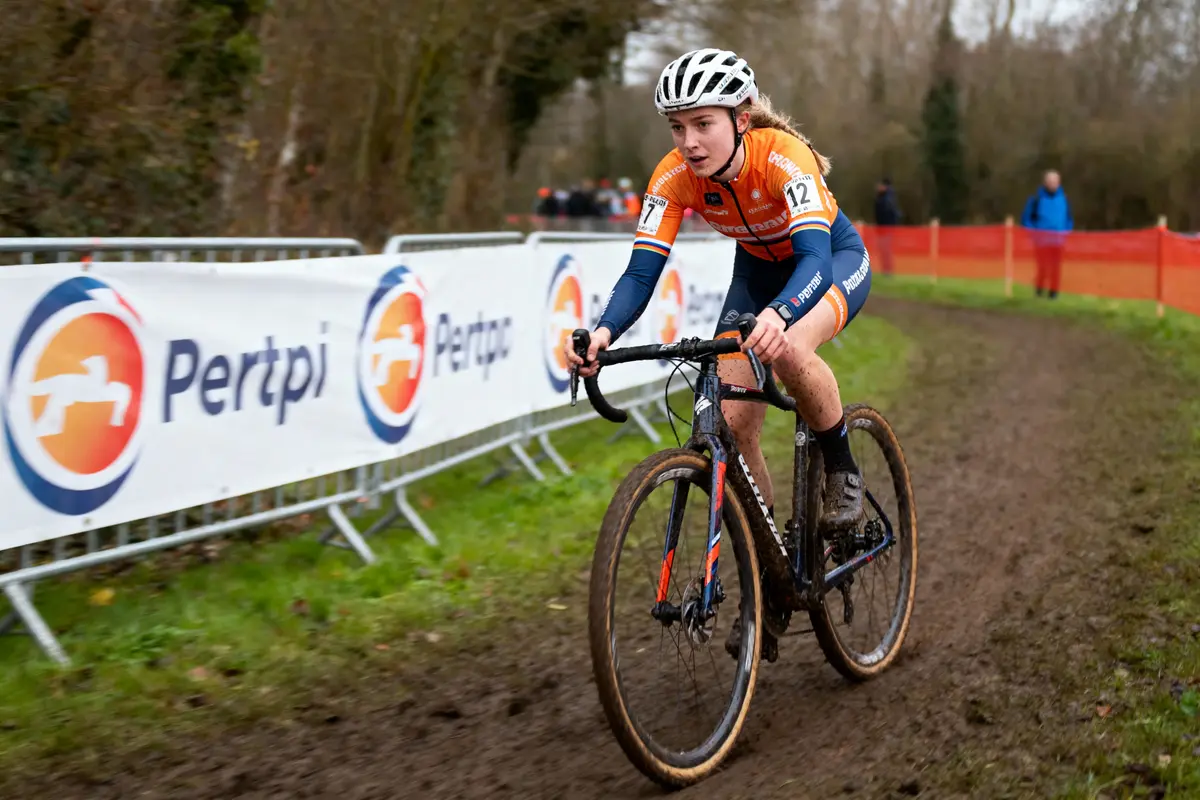 Cycliste en tenue orange participant à une course de cyclo-cross sur un chemin boueux, avec barrières publicitaires.