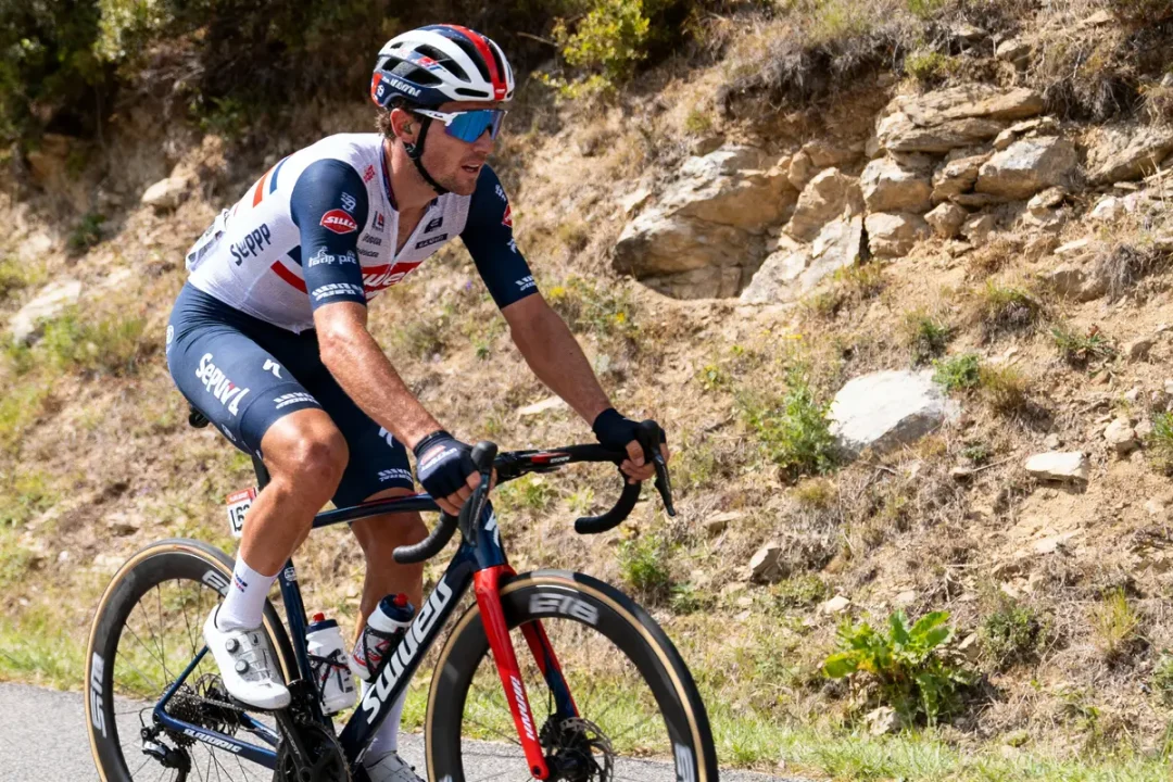 Cycliste en tenue professionnelle, roulant sur une route de montagne ensoleillée, entouré de végétation et rochers.