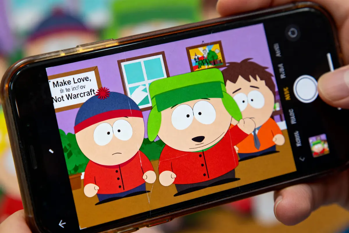 Personnages de South Park affichés sur un smartphone, avec un poster "Make Love, Not Warcraft" en arrière-plan.