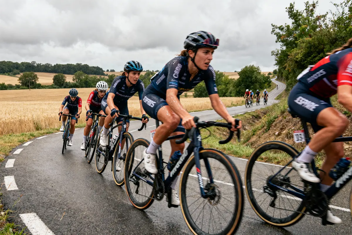 Cyclistes en compétition sur route sinueuse sous ciel nuageux, champs de blé en arrière-plan.