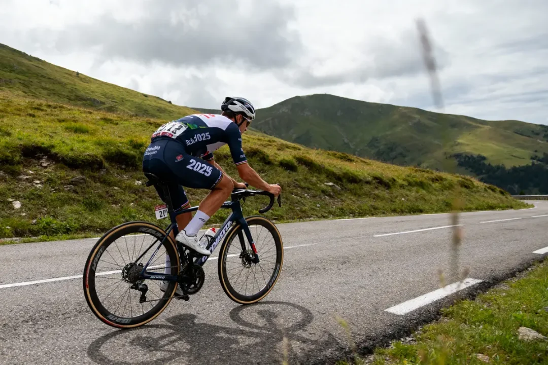 Cycliste en plein effort, sur route de montagne, ciel nuageux, paysage verdoyant.