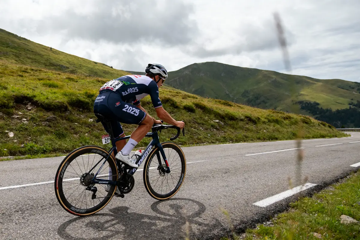 Cycliste en plein effort, sur route de montagne, ciel nuageux, paysage verdoyant.