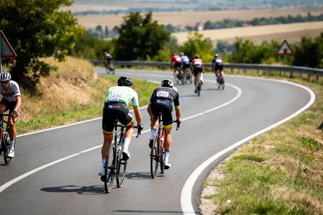Cyclistes participant à une course sur une route sinueuse, paysage rural en arrière-plan, ciel dégagé.