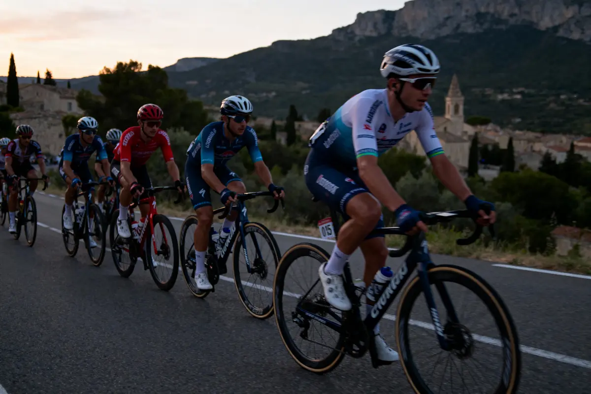 Cyclistes en course sur route montagneuse au crépuscule, église et collines en arrière-plan.