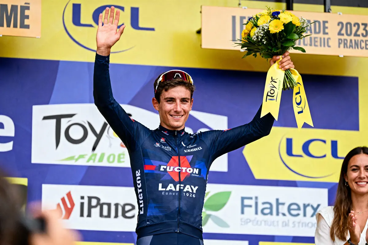 Cycliste en maillot bleu, levant la main, tenant un bouquet, sur un podium avec sponsor LCL en arrière-plan.