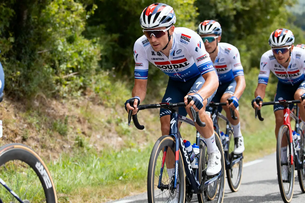 Cyclistes en tenue Soudal Quick-Step, roulant en équipe sur une route bordée de verdure.
