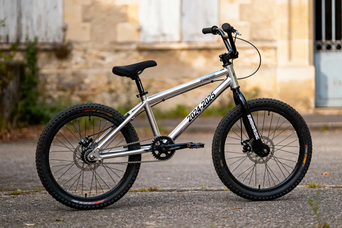 Vélo BMX argenté avec inscription "2024-2025" stationné devant un mur de pierre.