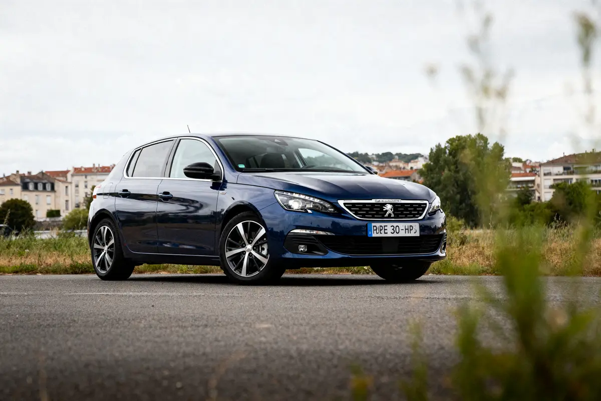 Voiture Peugeot bleue garée sur une route, bâtiments en arrière-plan, ciel nuageux.
