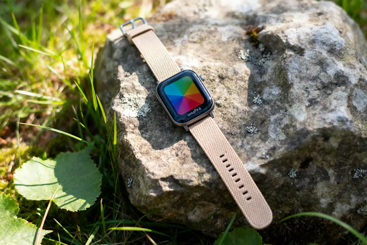 Montre connectée avec bracelet beige posée sur une pierre, entourée d'herbes et de feuilles vertes.