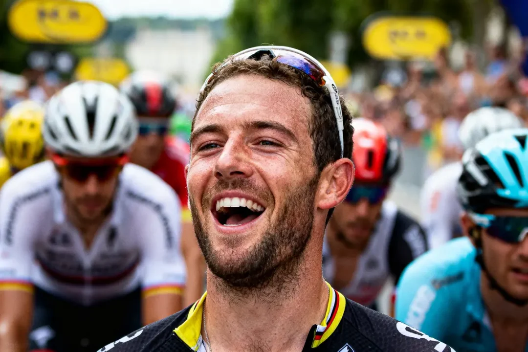 Cycliste souriant portant un maillot noir et lunettes, entouré d'autres coureurs lors d'une course, ambiance festive.