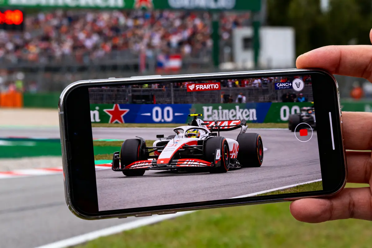 Smartphone filme une voiture de course en piste pendant une compétition, spectateurs flous en arrière-plan.