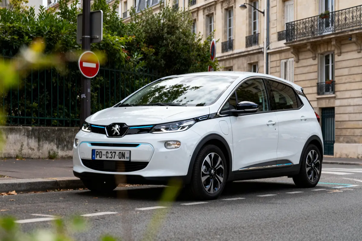Voiture Peugeot blanche garée en ville devant bâtiments et panneaux de signalisation.