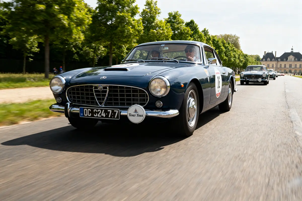 Voiture vintage noire roulant sur une route bordée d'arbres avec un château en arrière-plan.