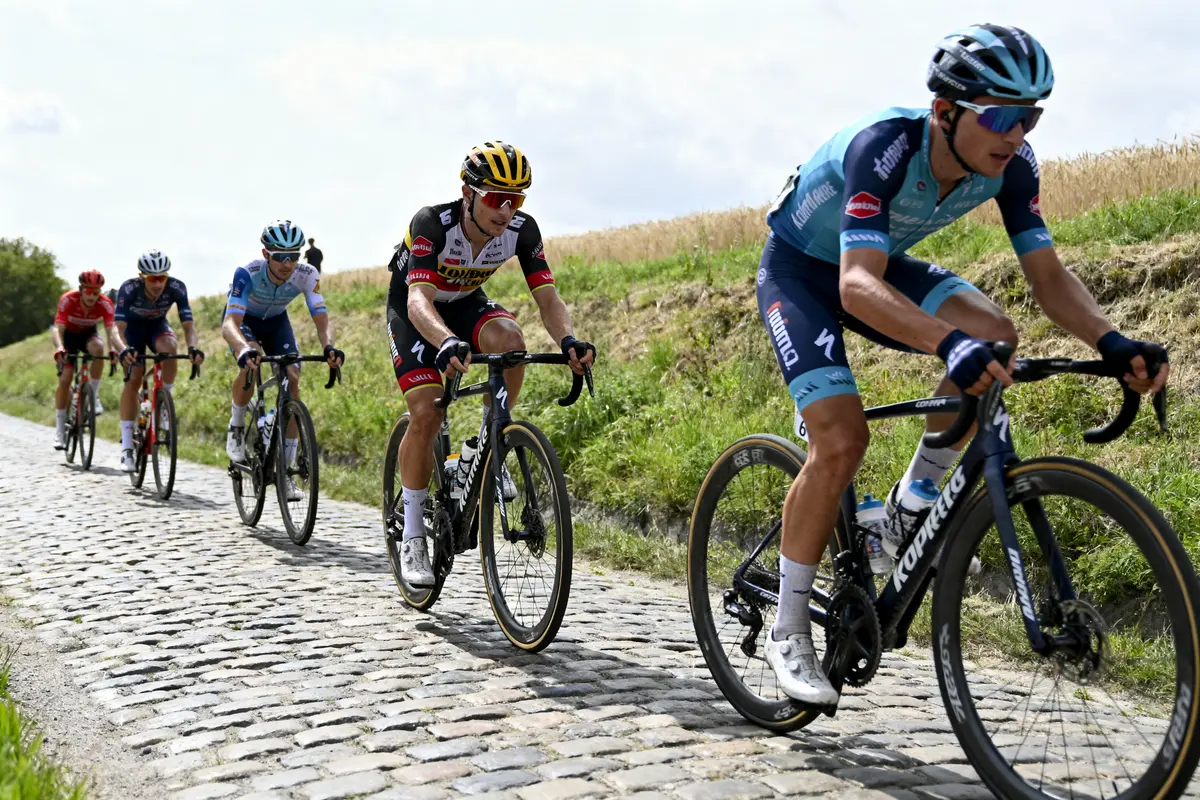 Cyclistes en course sur route pavée, campagne verdoyante, ciel légèrement nuageux, équipés de combinaisons colorées et casques.