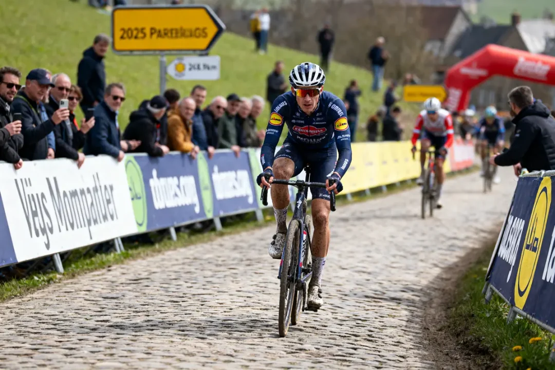 Cycliste sur route pavée, entouré de spectateurs, lors d'une course sous un ciel dégagé.