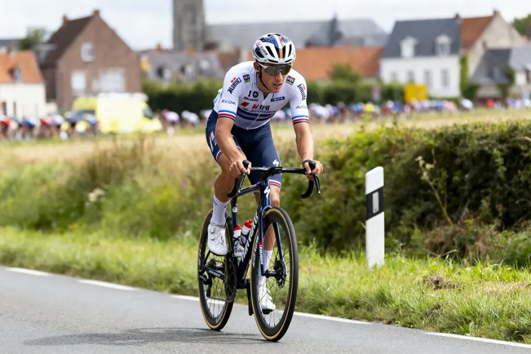 Cycliste en tenue blanche et bleue roulant sur une route de campagne, bâtiments en arrière-plan.