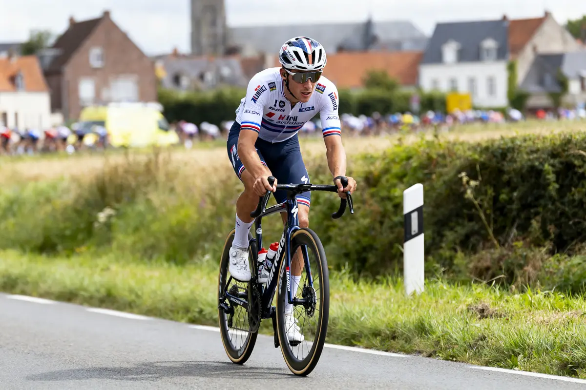 Cycliste en tenue blanche et bleue roulant sur une route de campagne, bâtiments en arrière-plan.