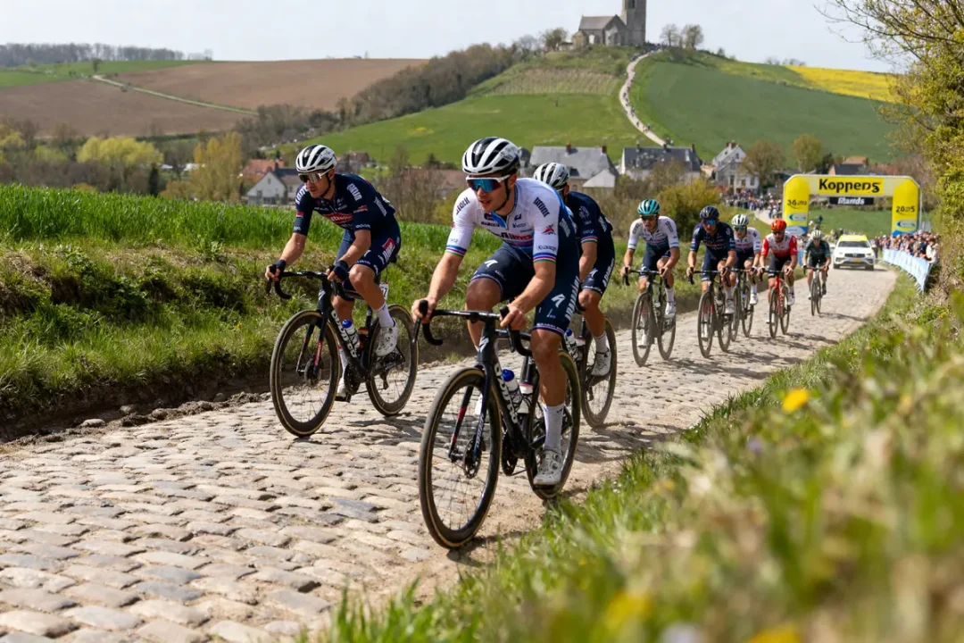 Cyclistes professionnels sur route pavée, paysage champêtre. Église au loin, ciel dégagé, course cycliste en cours.