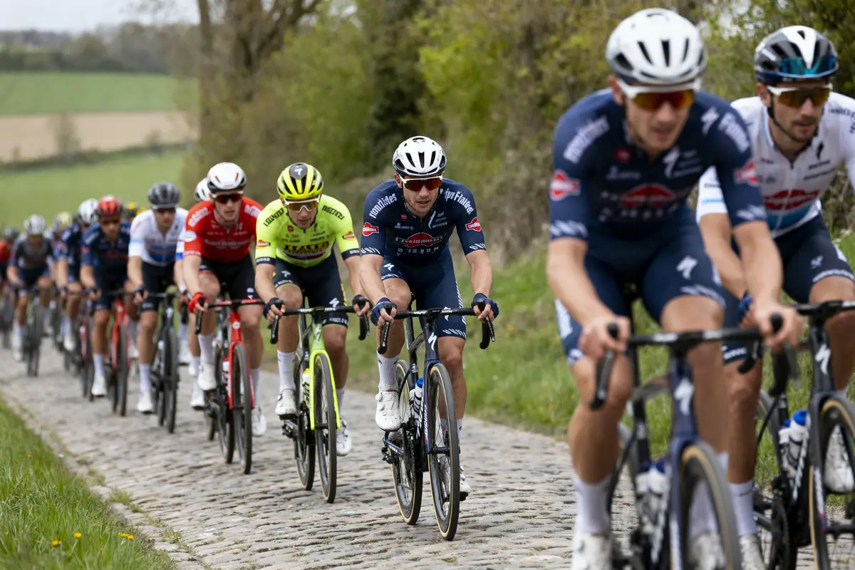 Cyclistes professionnels en course sur route pavée, portant divers maillots colorés, en pleine campagne.
