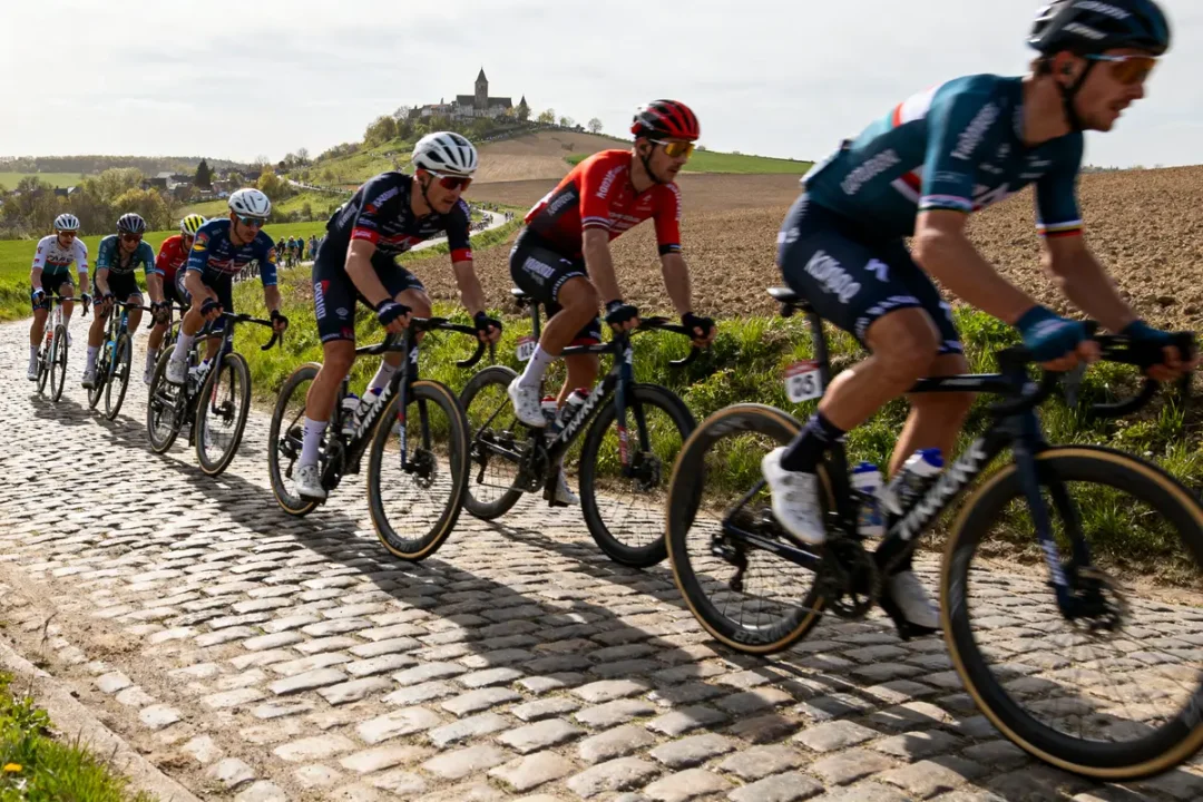 Cyclistes en course sur route pavée avec colline et village en arrière-plan. Conditions ensoleillées.