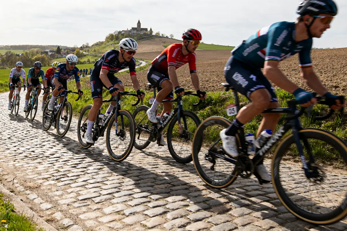 Cyclistes en course sur route pavée avec colline et village en arrière-plan. Conditions ensoleillées.