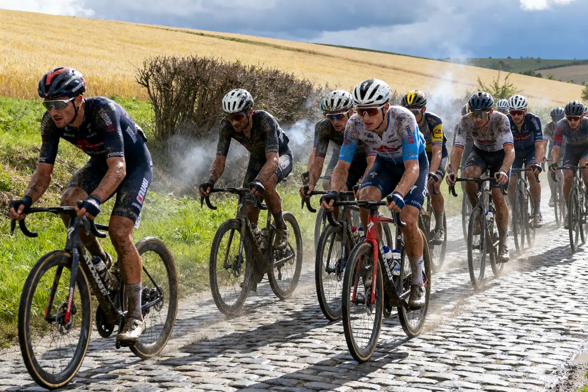 Cyclistes boueux roulant sur des pavés mouillés sous un ciel nuageux, champs en arrière-plan.