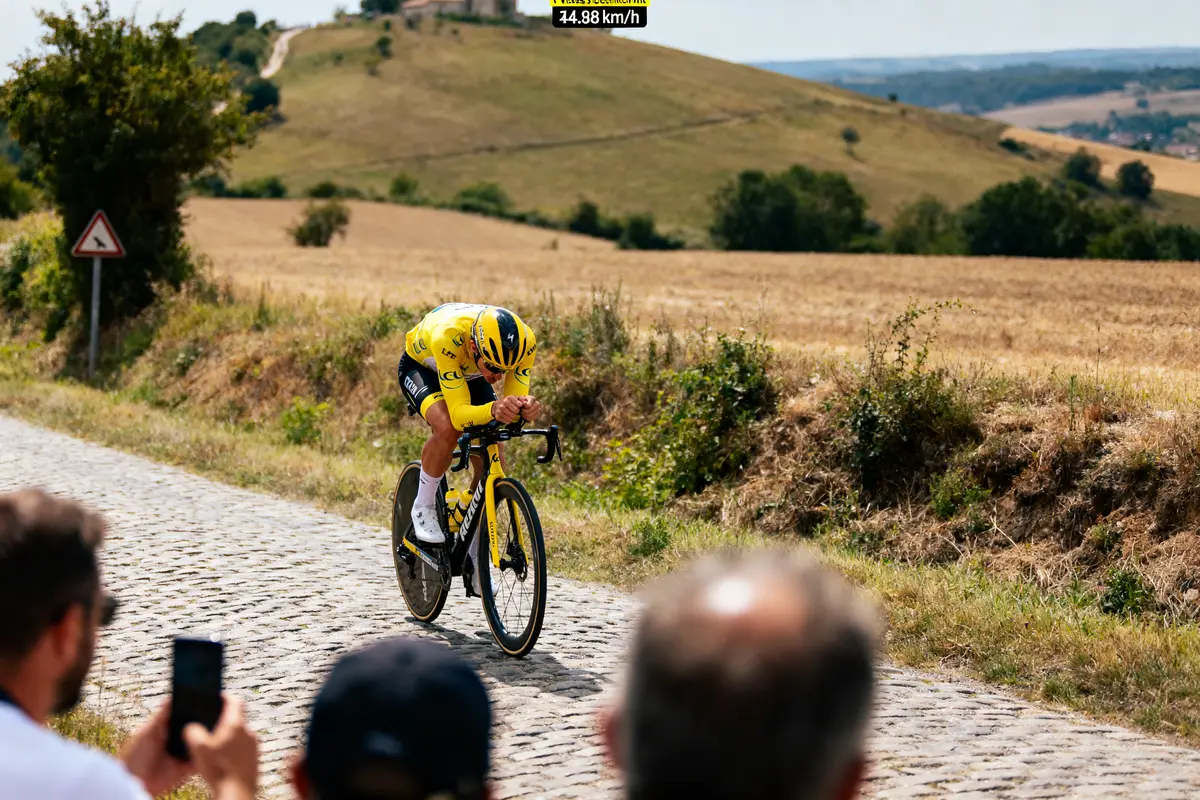 Cycliste en jaune sur route pavée, paysage rural, spectateurs au premier plan.