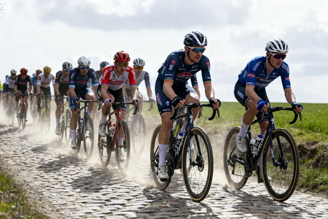 Cyclistes sur route pavée lors d'une course, spectateurs sur les côtés, paysage rural en arrière-plan.