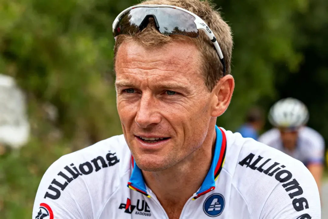 Cycliste en maillot blanc, lunettes de sport, avec fond flou de végétation verte.