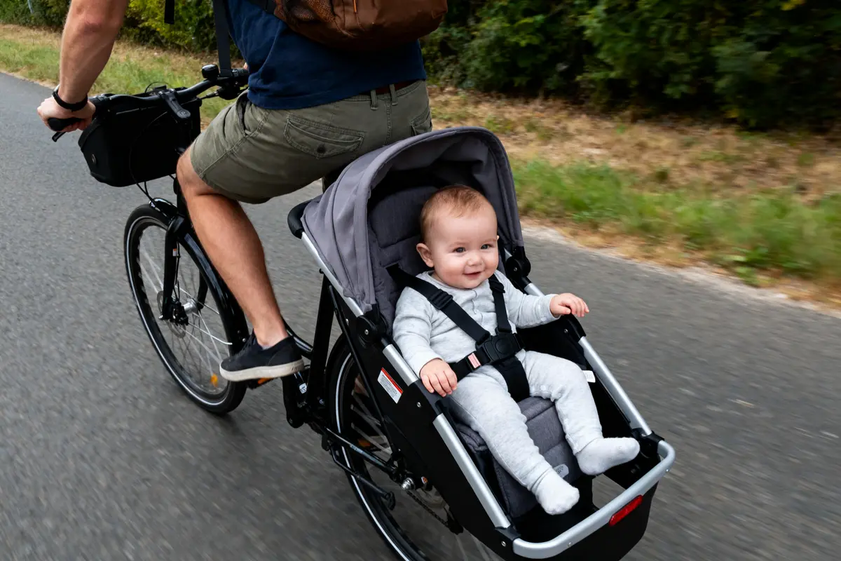 Un homme à vélo transporte un bébé souriant dans un siège remorque sur la route, verdure en arrière-plan.