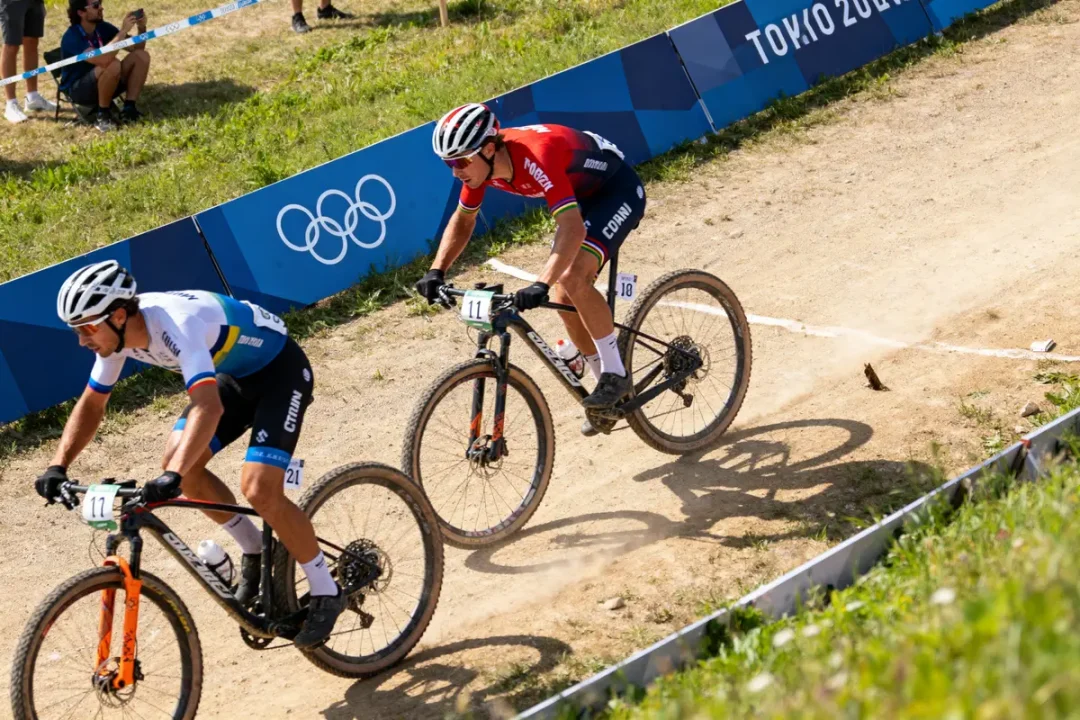 Deux cyclistes en compétition sur piste de VTT, devant un panneau Tokyo 2020 et logo Olympique.