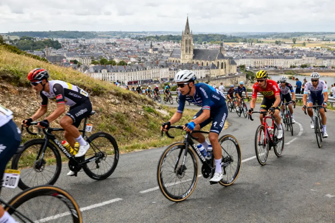 Cyclistes sur route vallonnée, Tour de France 2025 rumeurs, avec église et ville en fond