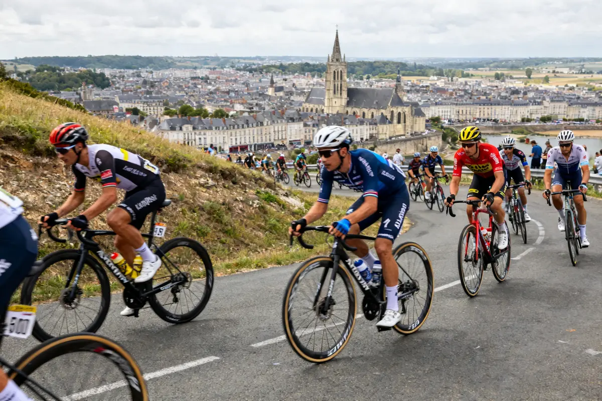 Cyclistes sur route vallonnée, Tour de France 2025 rumeurs, avec église et ville en fond